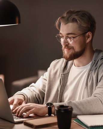 Ein junger Mann mit Brille sitzt an einem Tisch und arbeitet an einem Laptop. Er trägt einen grauen Hoodie und hat eine Tasse Kaffee sowie Notizmaterial neben sich. Die Beleuchtung ist gedämpft.