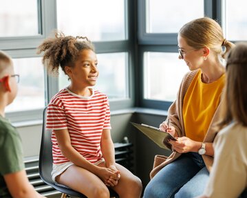 Ein Mädchen mit lockigem Haar in einem gestreiften Shirt sitzt auf einem Stuhl und spricht mit einer Frau, die Notizen macht. Zwei Kinder beobachten.