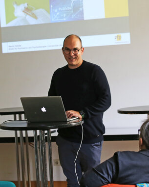 Ein Mann steht an einem Tisch mit einem Laptop und präsentiert vor einem Publikum in einem hellen Raum.