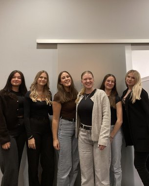 Sechs Frauen stehen in einer Reihe, lächelnd, in modischer Kleidung. Der Hintergrund zeigt eine einfache Wand und eine Glastür.