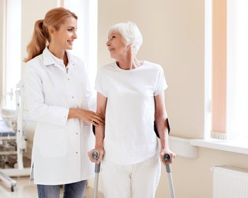 Eine Klinikmitarbeiterin unterstützt eine ältere Patientin, die mit Gehhilfen steht. Beide lächeln sich an, während sie in einem hellen Raum stehen.