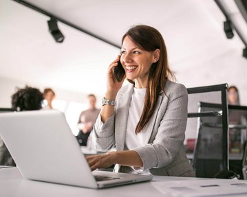Eine Frau spricht am Telefon, während sie an einem Laptop arbeitet. Im Hintergrund sind mehrere Personen in einem modernen Büro zu sehen.