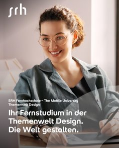 Eine lächelnde Frau mit Brille sitzt an einem Tisch und arbeitet an einem Laptop. Sie trägt ein graues Oberteil und hat lockige Haare. Im Hintergrund sind Fenster und ein helles Raumambiente sichtbar.