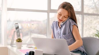 Eine Frau sitzt an einem Schreibtisch, spricht am Telefon und arbeitet an einem Laptop. Auf dem Tisch befinden sich Schreibmaterialien und eine kleine Pflanze.