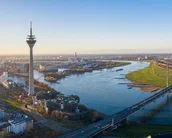 Der Rheinturm in Düsseldorf erhebt sich über die Stadt, umgeben von der Rheinpromenade und modernen Gebäuden. Im Hintergrund fließt der Rhein, während die Stadtlandschaft im Sonnenlicht erstrahlt.