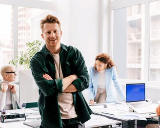 Ein Mann mit lässiger Kleidung steht lächelnd im Vordergrund eines modernen Büros, während zwei Personen im Hintergrund an einem Tisch arbeiten. Eine Frau beugt sich über einen Computer, während ein Mann am Tisch sitzt.
