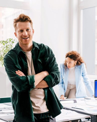 Ein Mann mit lässiger Kleidung steht lächelnd im Vordergrund eines modernen Büros, während zwei Personen im Hintergrund an einem Tisch arbeiten. Eine Frau beugt sich über einen Computer, während ein Mann am Tisch sitzt.