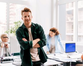 Ein Mann mit lässiger Kleidung steht lächelnd im Vordergrund eines modernen Büros, während zwei Personen im Hintergrund an einem Tisch arbeiten. Eine Frau beugt sich über einen Computer, während ein Mann am Tisch sitzt.