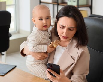 Eine Frau sitzt am Schreibtisch und hält ein Baby in den Armen, während sie auf ihr Smartphone schaut.
