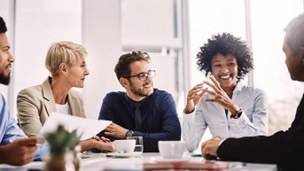 Eine Gruppe von fünf Personen sitzt an einem Tisch und diskutiert. Zwei Frauen und drei Männer zeigen Interesse und Engagement in der Unterhaltung, während sie Notizen und Getränke vor sich haben.