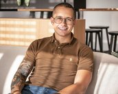 Ein junger Mann mit Brille sitzt auf einem Sofa. Er trägt ein braunes Polohemd und Jeans. Sein rechter Arm ist tätowiert. Im Hintergrund sind moderne Möbel und Pflanzen sichtbar.