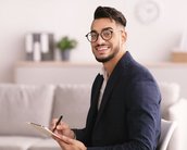Ein lächelnder Mann mit Brille sitzt auf einem Stuhl, hält einen Stift und ein Notizblatt in der Hand. Er trägt einen Anzug und befindet sich in einem modernen Raum mit einem Sofa im Hintergrund.