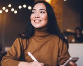 Eine lächelnde Frau mit langen, dunklen Haaren sitzt an einem Tisch, schreibt in ein Notizbuch und hält ein Smartphone in der Hand. Sie trägt einen braunen Pullover und wirkt konzentriert.
