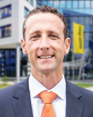 Ein Mann in einem Anzug mit orangefarbener Krawatte steht vor einem modernen Gebäude. Er lächelt und hat kurzes, lockiges Haar. Im Hintergrund ist ein gelbes Banner sichtbar.
