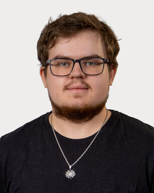 Ein junger Mann mit Brille und Bart trägt ein schwarzes T-Shirt und eine Halskette. Er schaut direkt in die Kamera.