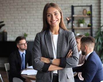 Eine lächelnde Frau in einem grauen Blazer steht im Vordergrund, während im Hintergrund mehrere Personen an einem Tisch in einem Besprechungsraum diskutieren.