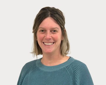 Eine lächelnde Person mit mittellangen, blonden Haaren trägt einen blauen Pullover. Der Hintergrund ist neutral und hell.