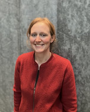 Eine Person mit blonden Haaren trägt eine rote Jacke und lächelt vor einer grauen Wand.