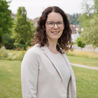 Eine Frau mit lockigem, braunem Haar trägt einen hellen Blazer und steht lächelnd im Freien. Im Hintergrund sind Bäume und eine Wiese sichtbar.