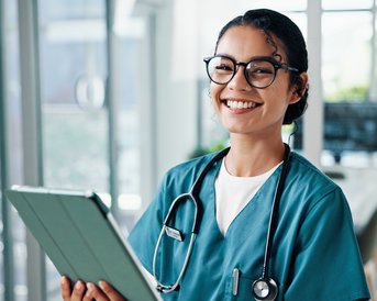 Eine lächelnde Krankenschwester in grüner Arbeitskleidung hält ein Tablet in der Hand. Im Hintergrund sind weitere medizinische Fachkräfte zu sehen.