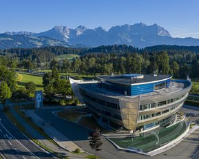 Moderne Architektur eines Gebäudes mit geschwungener Fassade, umgeben von grünen Wiesen und Bäumen, im Hintergrund eine Berglandschaft mit hohen Gipfeln und klarem Himmel.