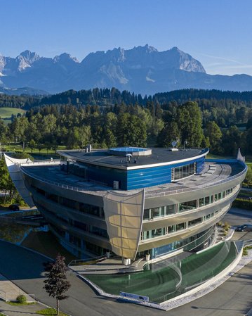 Moderne Architektur eines Gebäudes mit geschwungener Fassade, umgeben von grünen Wiesen und Bäumen, im Hintergrund eine Berglandschaft mit hohen Gipfeln und klarem Himmel.