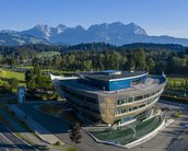 Moderne Architektur eines Gebäudes mit geschwungener Fassade, umgeben von grünen Wiesen und Bäumen, im Hintergrund eine Berglandschaft mit hohen Gipfeln und klarem Himmel.