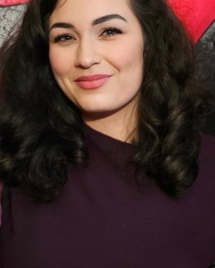 Eine Frau mit lockigem, dunklem Haar und einem lila Oberteil lächelt in die Kamera, vor einem bunten Hintergrund.