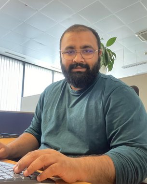 Ein Mann mit Bart sitzt an einem Schreibtisch und tippt auf einer Tastatur. Er trägt ein grünes Sweatshirt und eine Brille.
