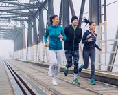 Drei Personen joggen auf einer Holzbrücke. Sie tragen sportliche Kleidung und scheinen aktiv und konzentriert zu sein. Die Umgebung ist urban und leicht neblig.