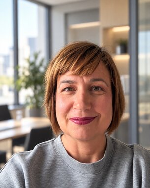 Frau mit schulterlangen, braunen Haaren und lila Lippenstift, lächelt in einem modernen Büro mit Pflanzen im Hintergrund.