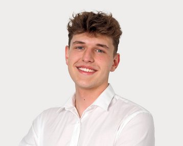 Junger Mann mit lockigem Haar, der in einem weißen Hemd lächelt und die Arme verschränkt. Neutraler Hintergrund.