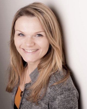 Eine lächelnde Frau mit langen, blonden Haaren steht vor einer hellen Wand. Sie trägt eine graue Jacke und einen orangefarbenen Oberteil.