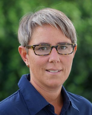 Eine Person mit kurzen, grauen Haaren und einer blauen Bluse trägt eine Brille mit einem tortoiseshell Muster. Der Hintergrund zeigt unscharfe grüne Pflanzen.