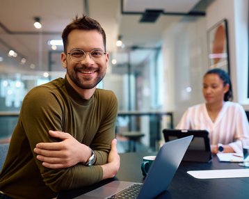 Ein Mann mit Brille lächelt am Tisch, während eine Frau im Hintergrund an einem Tablet arbeitet. Beide sind in einem modernen Büro.