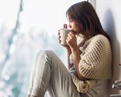 Eine Person sitzt am Fenster, trägt einen grob gestrickten Pullover und gemütliche Socken, während sie eine Tasse hält und daran nippt. Tageslicht strömt durch das Fenster.