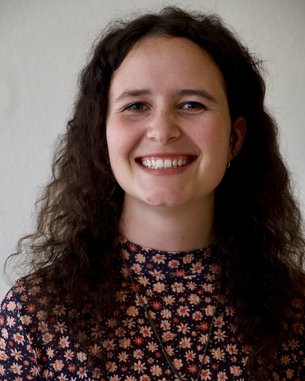Eine lächelnde Frau mit lockigem, braunem Haar trägt ein blumiges Oberteil und eine Halskette mit einem Symbol. Der Hintergrund ist neutral und hell.