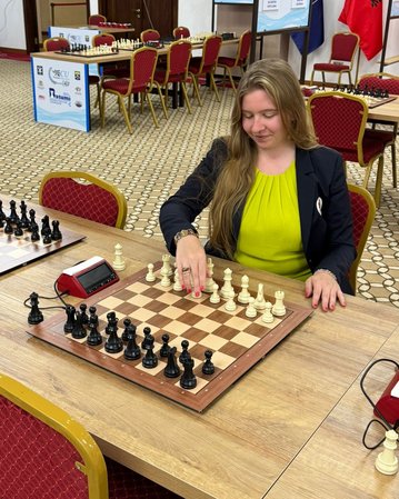 Eine Schachspielerin mit langen Haaren in einem schwarzen Blazer und gelbem Oberteil zieht eine Figur auf einem Schachbrett.