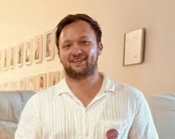 Ein Mann mit Bart trägt ein weißes Hemd und lächelt. Im Hintergrund sind gerahmte Porträts an der Wand zu sehen.