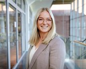 Eine lächelnde Frau mit blonden Haaren und Brille trägt einen karierten Blazer und steht in einem modernen Innenraum mit großen Fenstern.