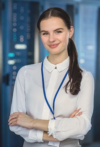 Eine junge Frau mit langen, dunklen Haaren steht in einem Serverraum, lächelt und trägt ein weißes Hemd mit einer blauen Halskette. Im Hintergrund sind Serverregale sichtbar.