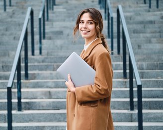 Junge Frau mit braunem Mantel steht an einer Treppe und hält einen Laptop in der Hand. Sie lächelt in die Kamera.
