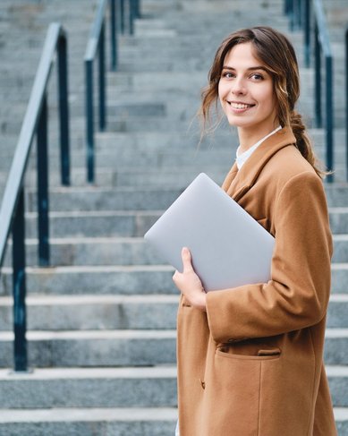 Junge Frau mit braunem Mantel steht an einer Treppe und hält einen Laptop in der Hand. Sie lächelt in die Kamera.