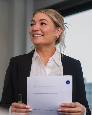 Eine Frau in einem schwarzen Blazer hält Dokumente in der Hand und spricht, während sie lächelt. Im Hintergrund sind Fenster sichtbar.