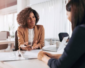 Eine Frau mit lockigem Haar trägt einen braunen Blazer und spricht mit einer anderen Person am Tisch. Auf dem Tisch liegen Dokumente und ein Laptop.