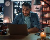 Ein Mann sitzt an einem Tisch und lächelt, während er auf einen Laptop schaut. Neben ihm stehen eine Tasse und eine Kerze. Im Hintergrund sind Bücherregale und Fenster mit Stadtansicht zu sehen.