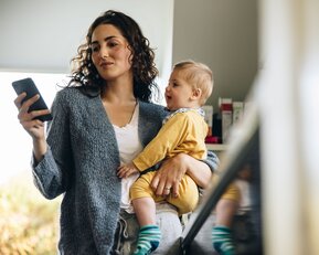 Eine Frau mit lockigem Haar hält ein Smartphone in der Hand und trägt ein Kleinkind in einem gelben Outfit.