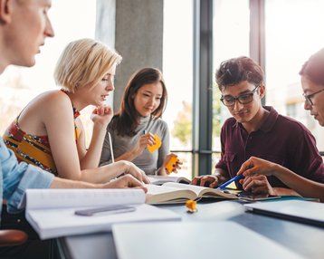 Sechs Personen sitzen an einem Tisch und diskutieren über ein Buch. Einige halten Notizen oder Stifte, während sie sich auf das Material konzentrieren. Tageslicht strömt durch große Fenster.