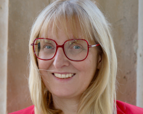 Eine Frau mit langen blonden Haaren und roten Brillenläsern lächelt freundlich. Sie trägt ein rotes Oberteil und steht vor einer neutralen Wand.