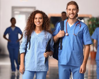 Zwei medizinische Fachkräfte in blauen Uniformen stehen lächelnd in einem Flur, bereit für ihre Aufgaben.